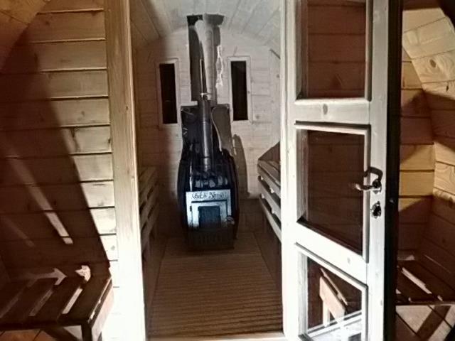 Wnętrze drewnianej sauny z piecem, z widocznym dymem z komina, częściowo otwarte białe drzwi z drewnianymi wstawkami.