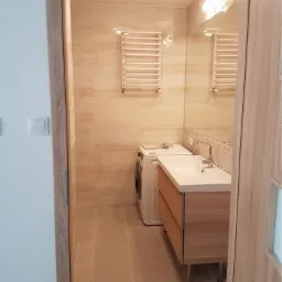 Wykończona łazienka w beżowych odcieniach z drewnianą szafką pod umywalką, pralką i grzejnikiem drabinkowym, widok zza otwartych drzwi.