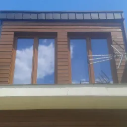 Fragment elewacji budynku z drewnianą okładziną i dwoma oknami, w których odbija się błękitne niebo z chmurami; widoczna antena satelitarna na ścianie.