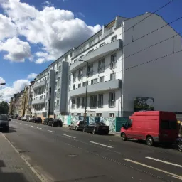 Nowoczesny, trzypiętrowy budynek mieszkalny z balkonami, pomalowany na biało z szarymi akcentami, widoczny z ulicy w słoneczny dzień, z zaparkowanymi samochodami i czerwonym vanem w pobliżu.