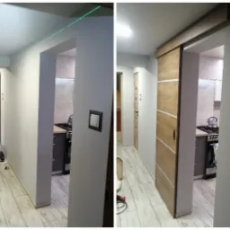 Montaż drewnianych drzwi przesuwnych w korytarzu, widoczny laser poziomujący na ścianie, częściowo wyposażona kuchnia w tle.