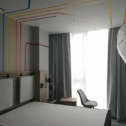 Pokój hotelowy z minimalistycznym wystrojem, łóżkiem bez pościeli, szarą zasłoną i dekoracją ścienną w postaci kolorowych linii imitujących instalacje.