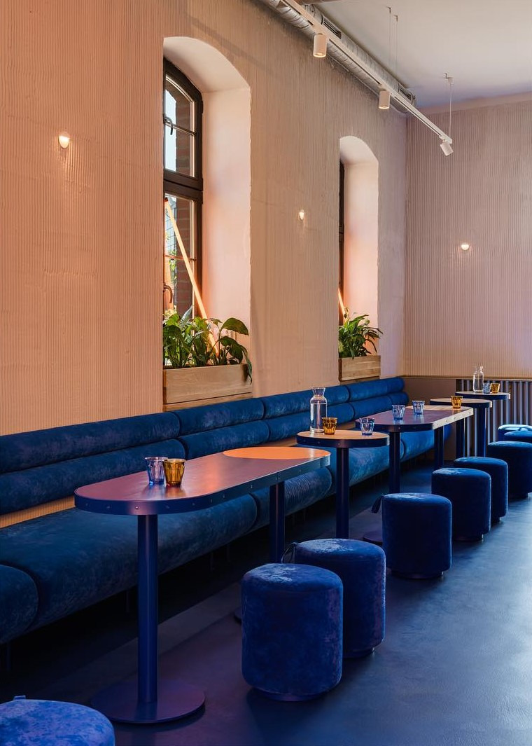 Wnętrze kawiarni z długą, niebieską, tapicerowaną ławą, okrągłymi stolikami z drewnianymi blatami i niebieskimi pufami. Na stolikach stoją szklanki i karafki z wodą. W tle okna z roślinami...