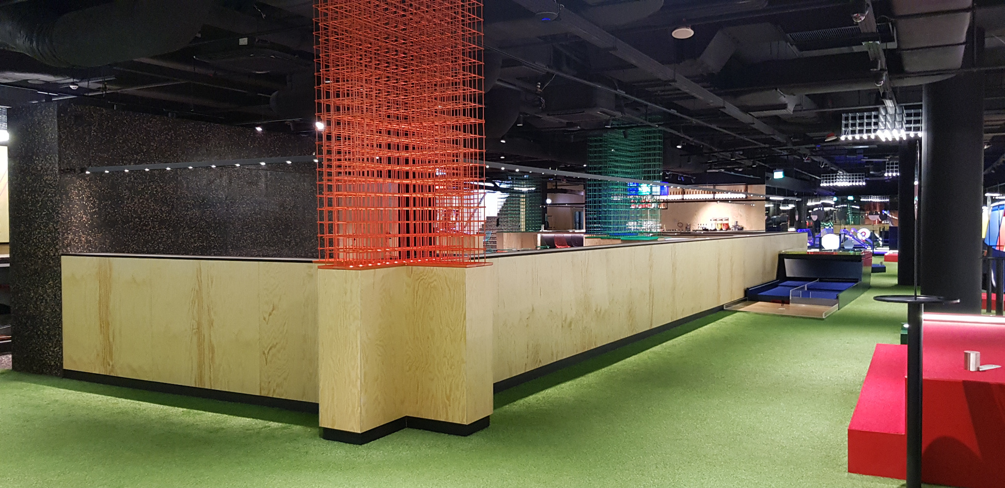 Długa, jasna lada z drewnianymi frontami w lokalu rozrywkowym, z elementami dekoracyjnymi w postaci czerwonej i zielonej kratownicy, na podłodze sztuczna trawa