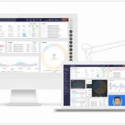 Ekran komputera stacjonarnego i laptopa prezentujące dashboard z wykresami i danymi finansowymi, oświetlone białą lampką biurową.
