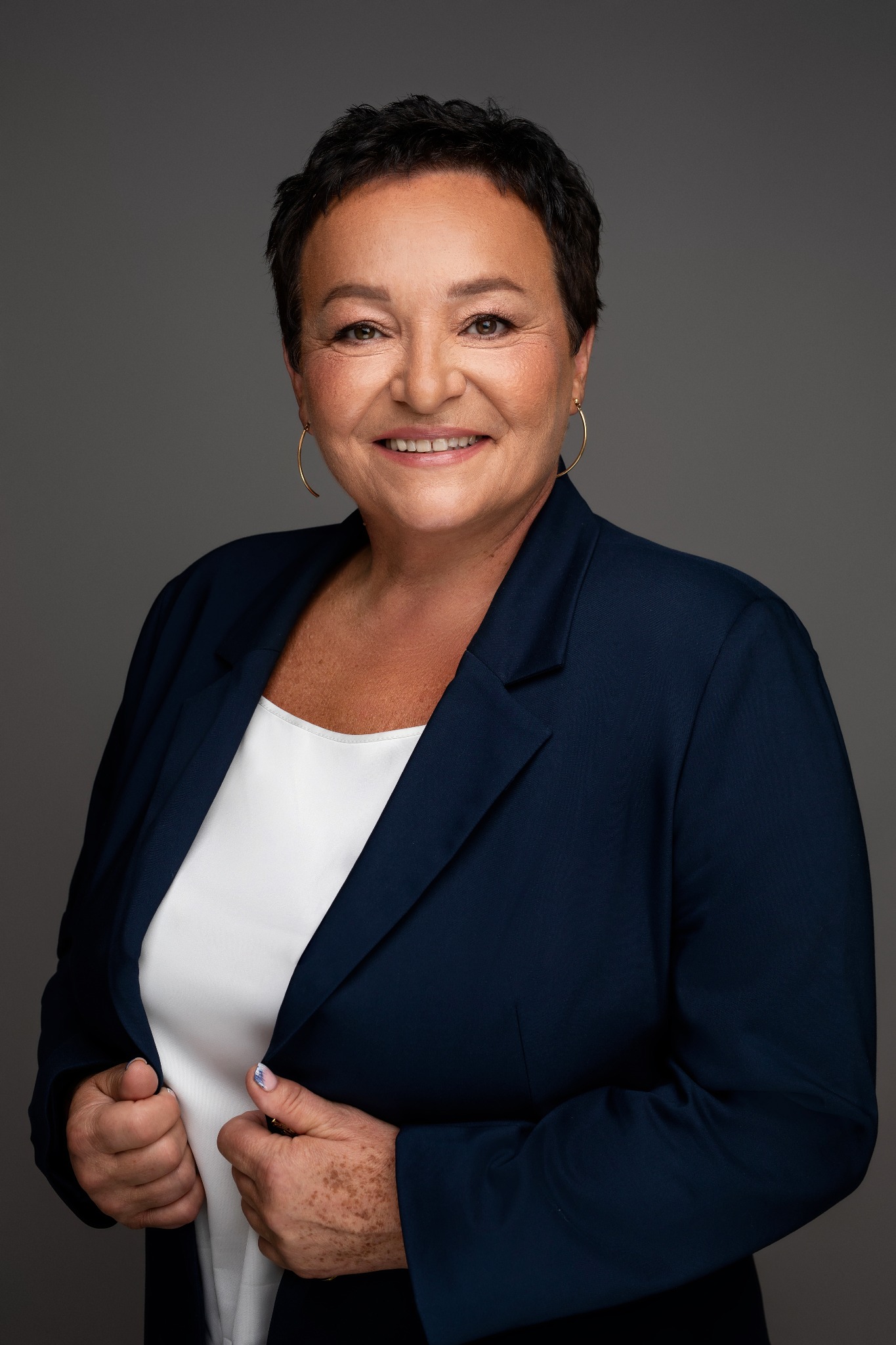 Portret uśmiechniętej kobiety w granatowej marynarce i białej bluzce, z krótkimi ciemnymi włosami i złotymi kolczykami na szarym tle.