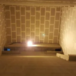 Wnętrze w trakcie prac wykończeniowych, ściany wyłożone płytami, widoczne łączenia płyt, oświetlenie robocze, surowe schody wiodące w dół.