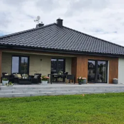 Nowoczesny dom jednorodzinny z tarasem, czarną dachówką i stolarką okienną w kolorze antracytu, widok z ogrodu w pochmurny dzień.