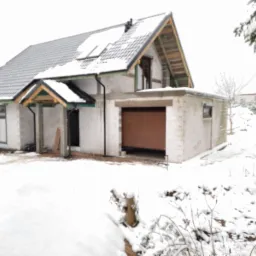 Niedokończony dom jednorodzinny w zimowej scenerii, widoczna drewniana brama garażowa w trakcie montażu, okna z brązowymi ramami, oraz ośnieżony dach pokryty szarą dachówką.