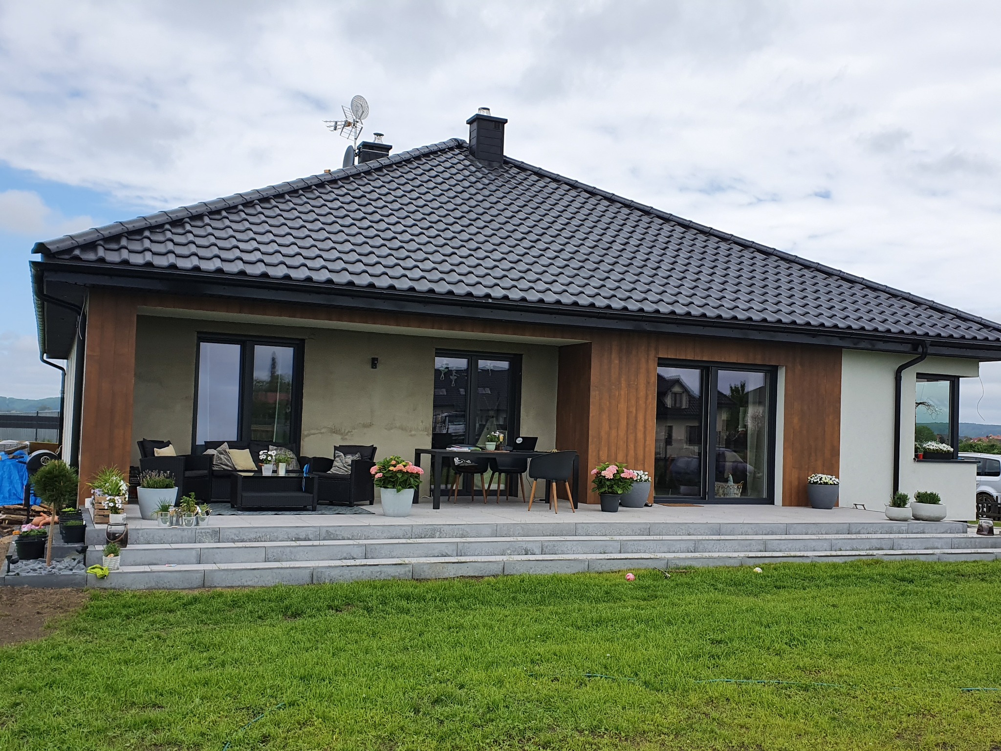 Nowoczesny dom jednorodzinny z tarasem, czarną dachówką i stolarką okienną w kolorze antracytu, widok z ogrodu w pochmurny dzień.