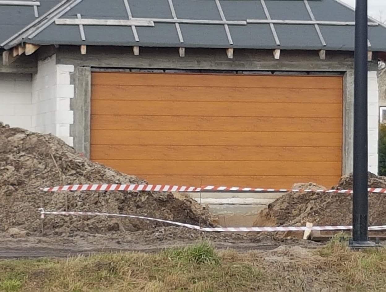 Nowa, drewnopodobna brama garażowa segmentowa w surowym stanie, widoczna ziemia i taśma ostrzegawcza.