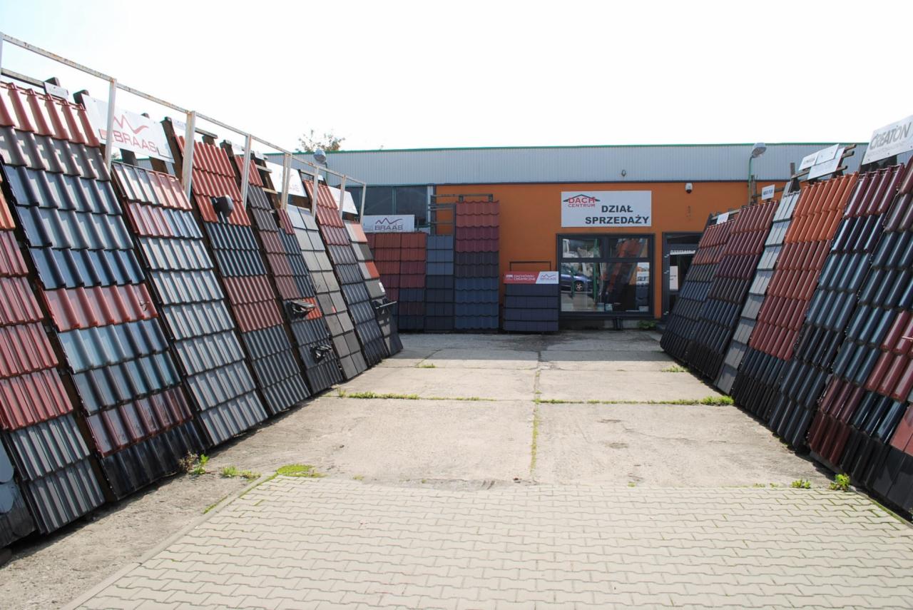 Ekspozycja różnych modeli i kolorów dachówek w Krakowie, ustawionych na stojakach przed budynkiem z napisem 'Dział Sprzedaży'. Widoczne logo producentów na niektórych modelach.
