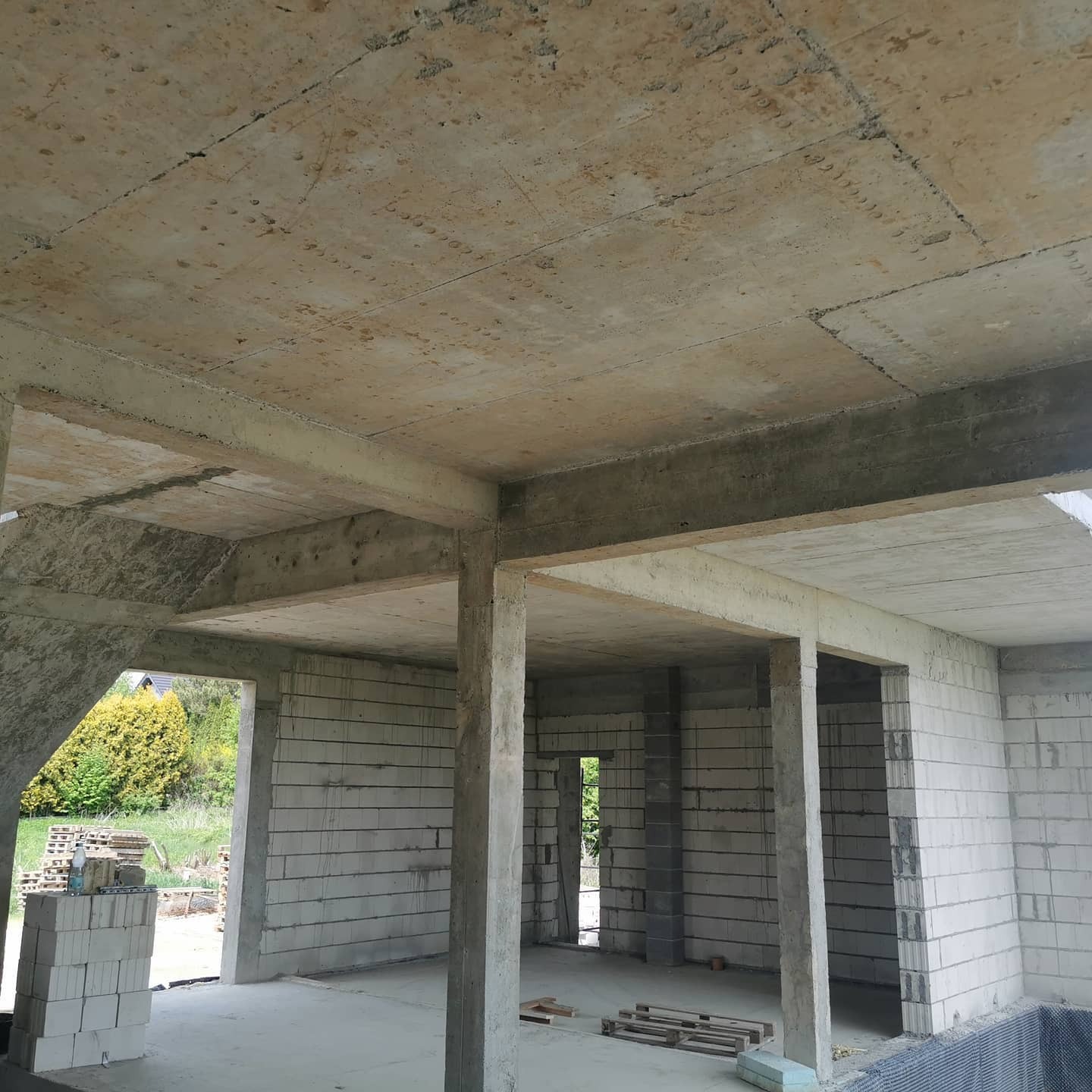 Surowy stan budynku w trakcie budowy: ściany z bloczków, betonowy strop z widocznymi śladami szalunków, kolumny podtrzymujące, widok na zieleń przez otwory okienne.