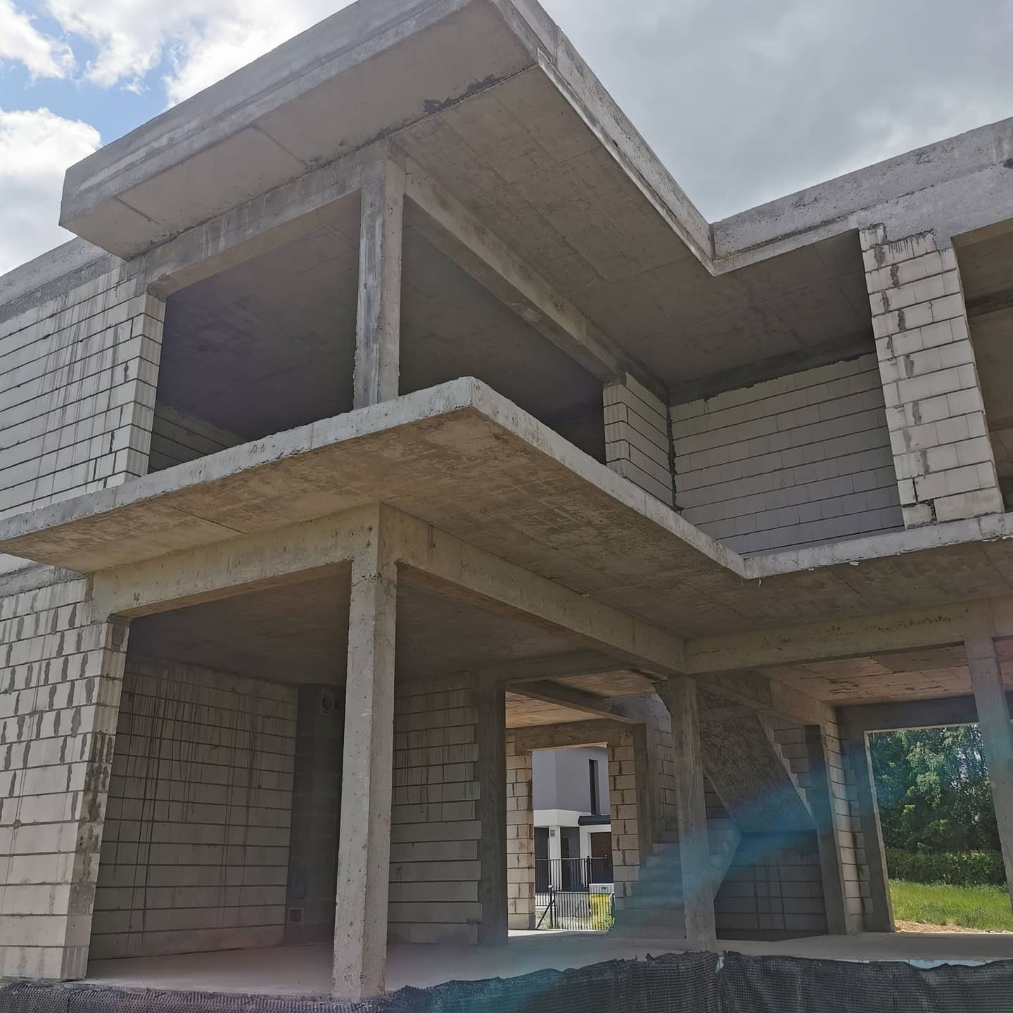 Surowy stan dwukondygnacyjnego budynku z betonu komórkowego, widoczne ściany, stropy i słupy konstrukcyjne, zarys okien i drzwi, w tle zieleń.