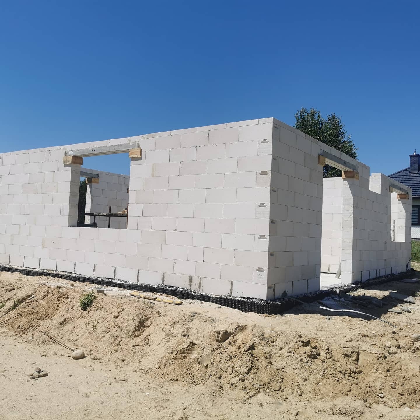 Wznoszone ściany parterowe domu z białych bloczków na fundamencie z izolacją przeciwwilgociową, widoczne nadproża okienne z drewnianych belek, piaszczysty teren budowy w słoneczny dzień.
