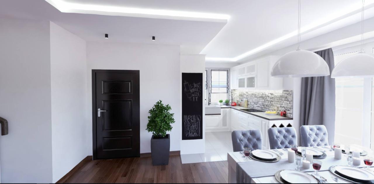 Nowoczesne wnętrze w Poznaniu: połączenie przedpokoju z kuchnią i jadalnią, białe ściany, drewniana podłoga, czarne drzwi, tablica kredowa z napisem, roślina w donicy, biała kuchnia z ceglaną...