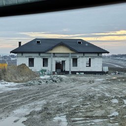 Dawid Wilk TYNK-TECH - Surowy stan domu z dachem w Koniuszy, widok zza szyby. Budynek w trakcie budowy, otoczony błotnistym terenem. Widoczne pustaki i elementy konstrukcyjne.