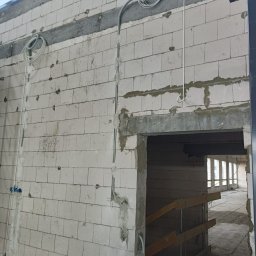 Dawid Wilk TYNK-TECH - Wewnętrzna ściana z betonu komórkowego w trakcie budowy, z widocznymi przewodami instalacji elektrycznej i puszkami pod gniazdka. Surowy stan, widok na kolejne pomieszczenie.