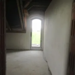 Wnętrze pomieszczenia w trakcie remontu: surowe ściany po nałożeniu warstwy tynku, widoczna instalacja elektryczna przy podłodze, okno z widokiem na zieleń, sufit z drewnianą konstrukcją.