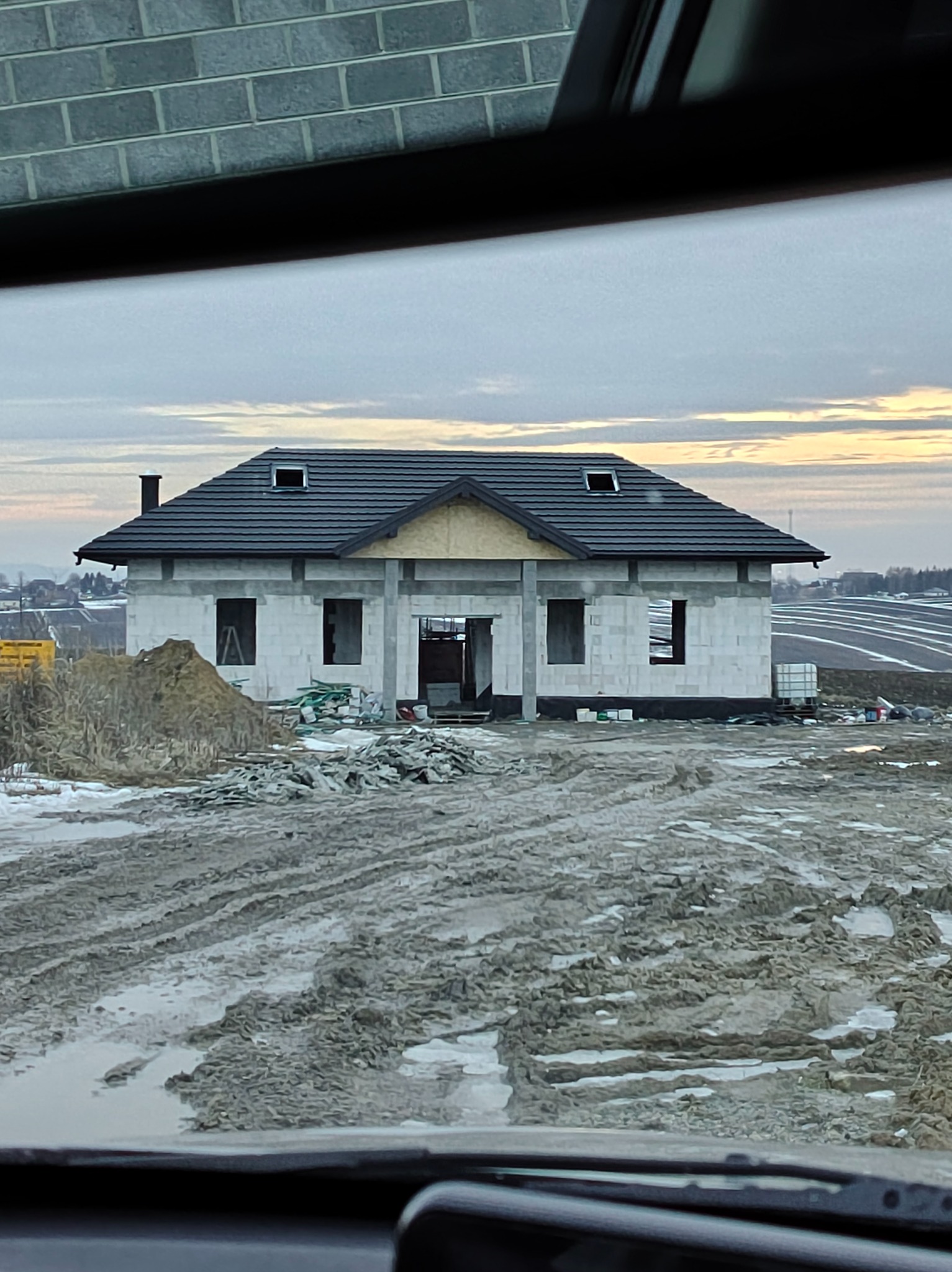 Surowy stan domu z dachem w Koniuszy, widok zza szyby. Budynek w trakcie budowy, otoczony błotnistym terenem. Widoczne pustaki i elementy konstrukcyjne.