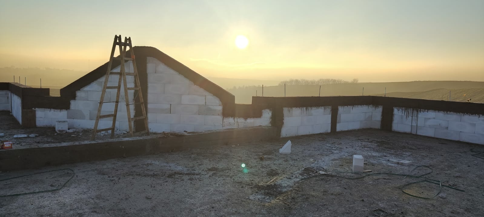 Wznoszenie ścian z bloczków betonu komórkowego na tle wschodzącego słońca. Widoczna drewniana drabina oparta o szczyt ściany. Etap budowy domu w promieniach porannego światła.