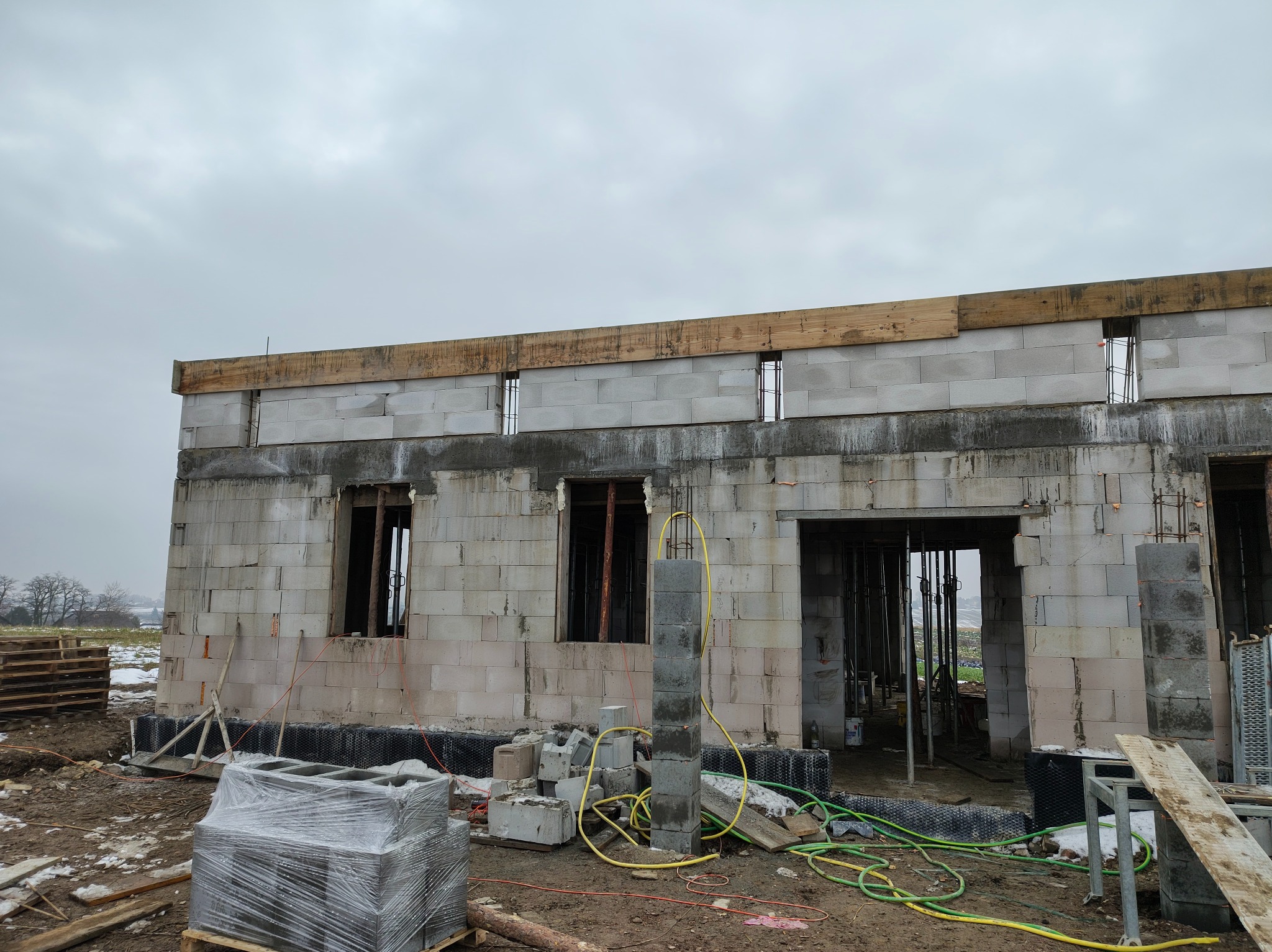 Budowa domu z betonu komórkowego w trakcie realizacji. Widoczne ściany, otwory okienne i drzwiowe, drewniane nadproża oraz materiały budowlane na placu budowy.