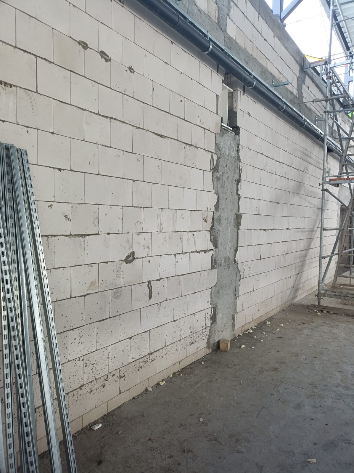 Ściana z betonu komórkowego z bruzdą wypełnioną zaprawą, widoczne rury instalacyjne pod sufitem i metalowe profile po lewej stronie. Budowa w trakcie realizacji.