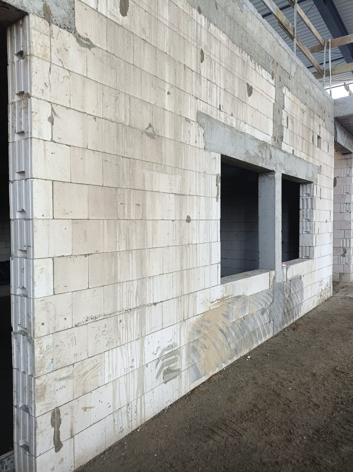 Ściana budynku w trakcie murowania z betonu komórkowego, widoczne otwory okienne z nadprożami. Ślady zaprawy na ścianie. Konstrukcja dachu w tle.