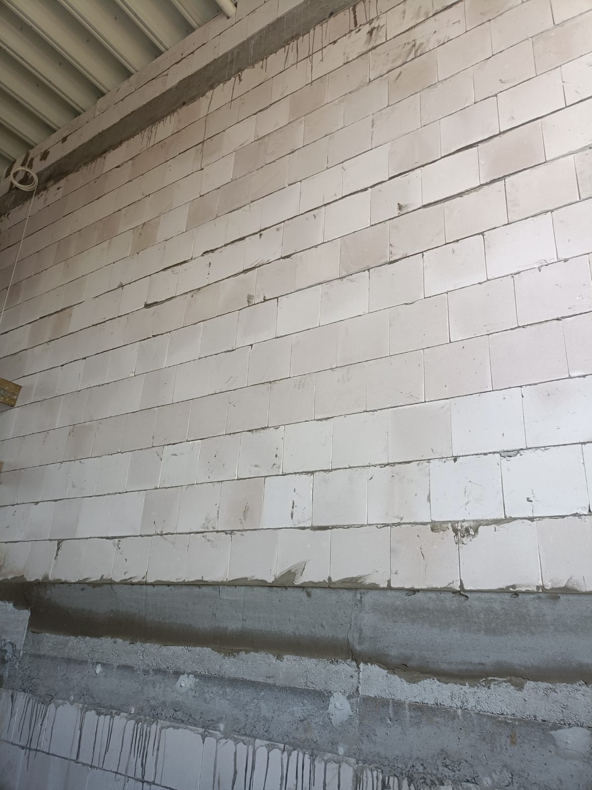 Ściana z białych bloczków, widoczne spoiny i fragment stropu. Dolna część ściany w surowym stanie, z widocznymi śladami zaprawy. Ujęcie z dołu, lekko pod kątem.
