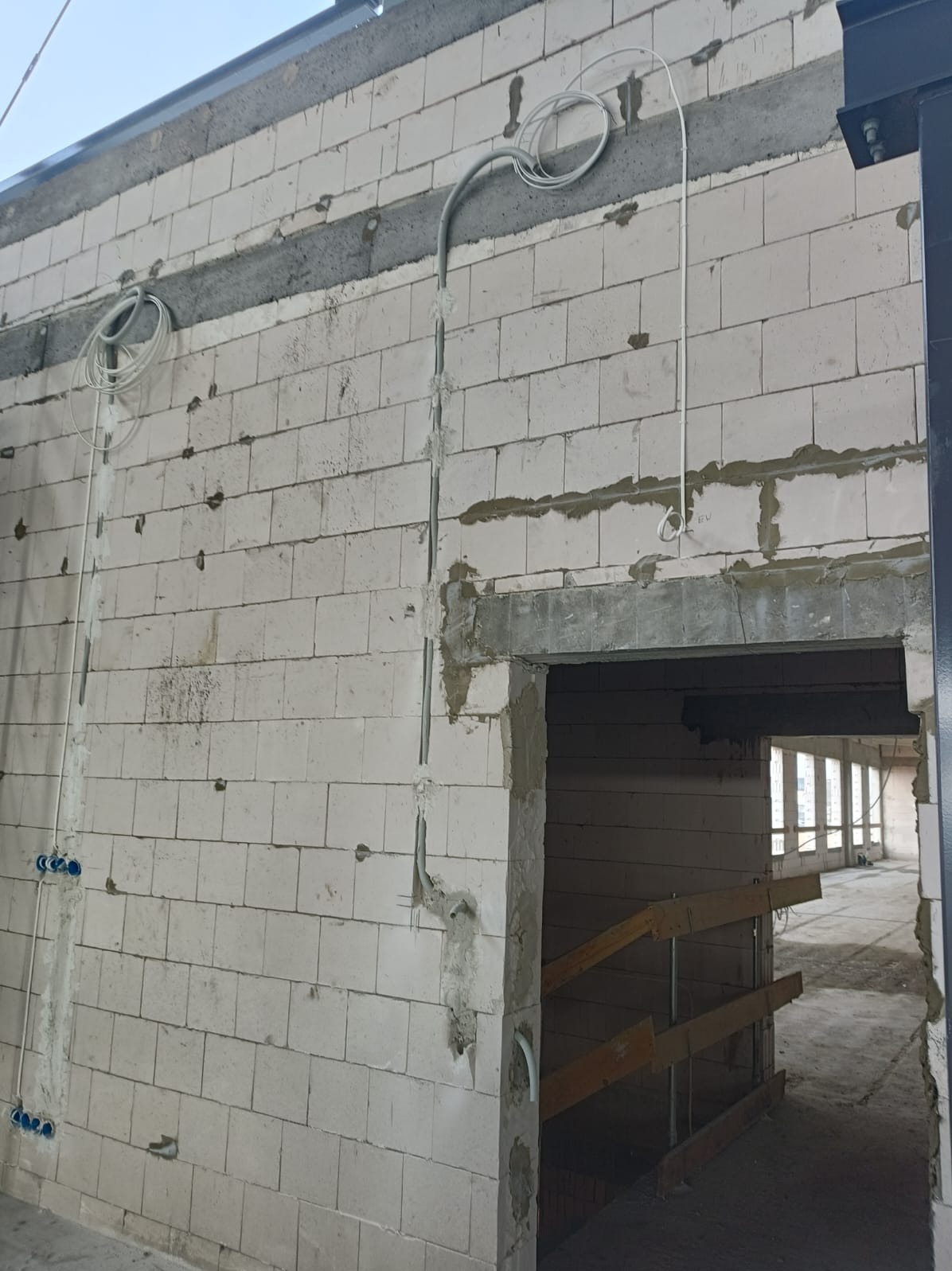 Wewnętrzna ściana z betonu komórkowego w trakcie budowy, z widocznymi przewodami instalacji elektrycznej i puszkami pod gniazdka. Surowy stan, widok na kolejne pomieszczenie.