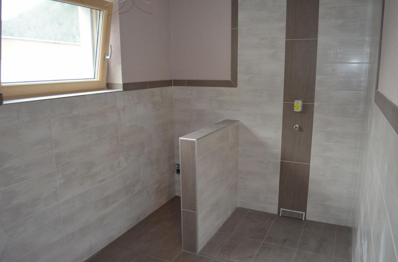 Nowoczesna kabina prysznicowa walk-in z beżowymi płytkami imitującymi drewno, z ciemnobrązowym pionowym pasem na ścianie, zakończona odpływem liniowym i małym okienkiem z drewnianą ramą.