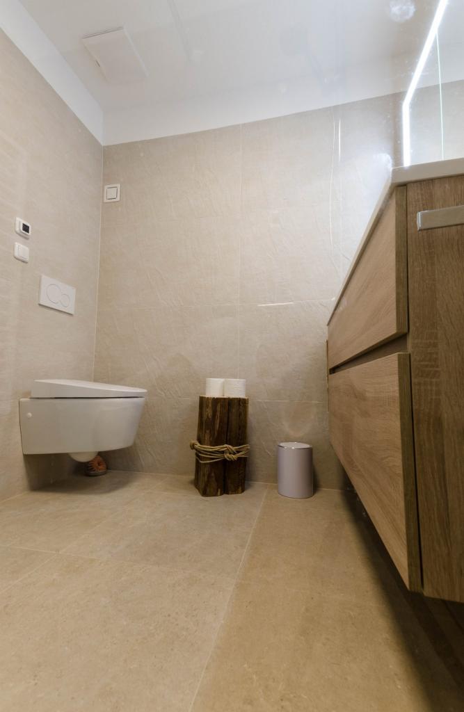 Nowoczesna łazienka z podwieszaną toaletą, beżowymi płytkami, drewnianą szafką pod umywalką i dekoracyjnym stojakiem na papier toaletowy z drewna.