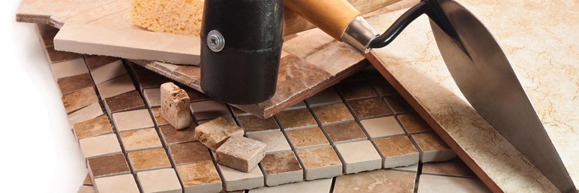 Mozaika ceramiczna w odcieniach beżu i brązu, czarny gumowy młotek, kielnia i gąbka do fugowania na jasnym tle.