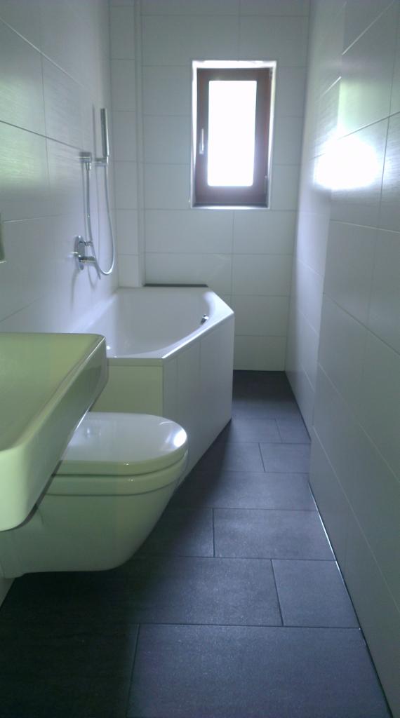 Wnętrze łazienki z białymi płytkami ściennymi, ciemnoszarą podłogą z płytek, białą wanną narożną i toaletą z umywalką, oświetlone naturalnym światłem z okna.