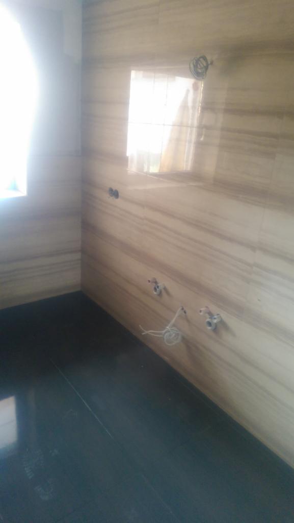 Wykończona łazienka z położoną ciemną podłogą i beżowymi płytkami imitującymi marmur na ścianach, widoczne wyprowadzenia instalacji wodnej i elektrycznej, przygotowane do montażu armatury.