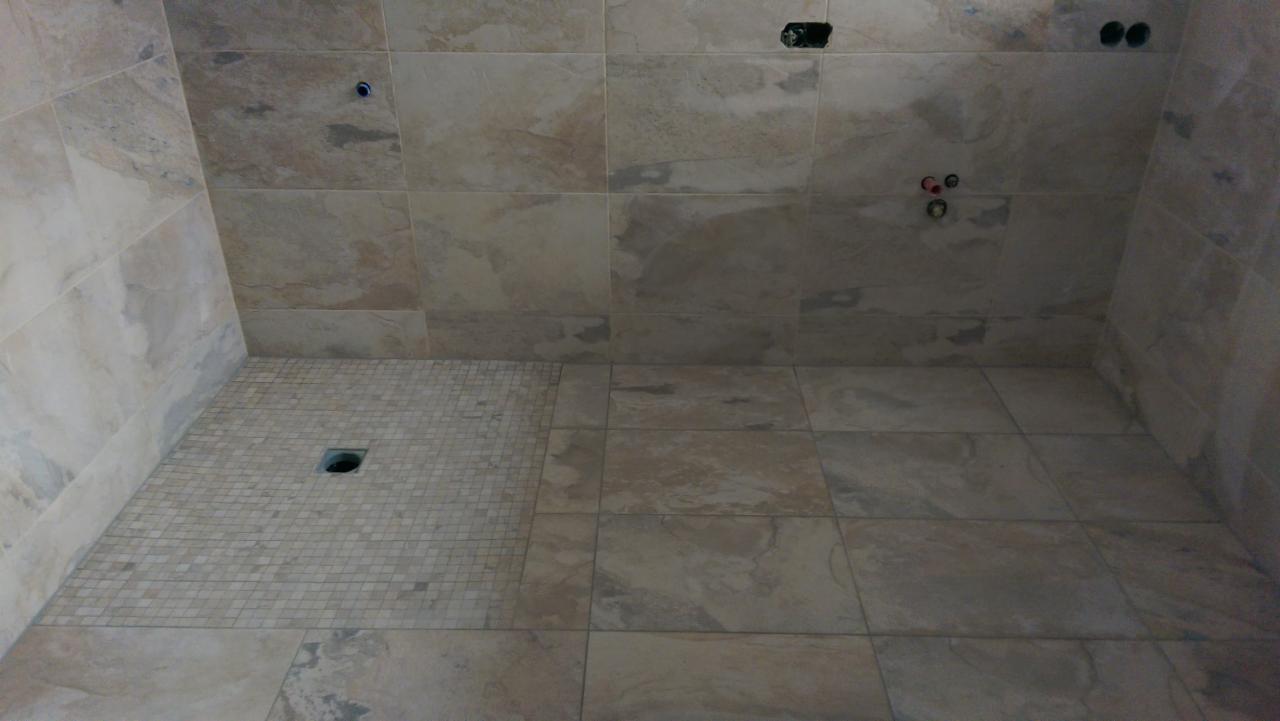 Wykończona kabina prysznicowa z beżowymi płytkami imitującymi kamień, z odpływem liniowym wyłożonym mozaiką i widocznymi otworami na instalacje.