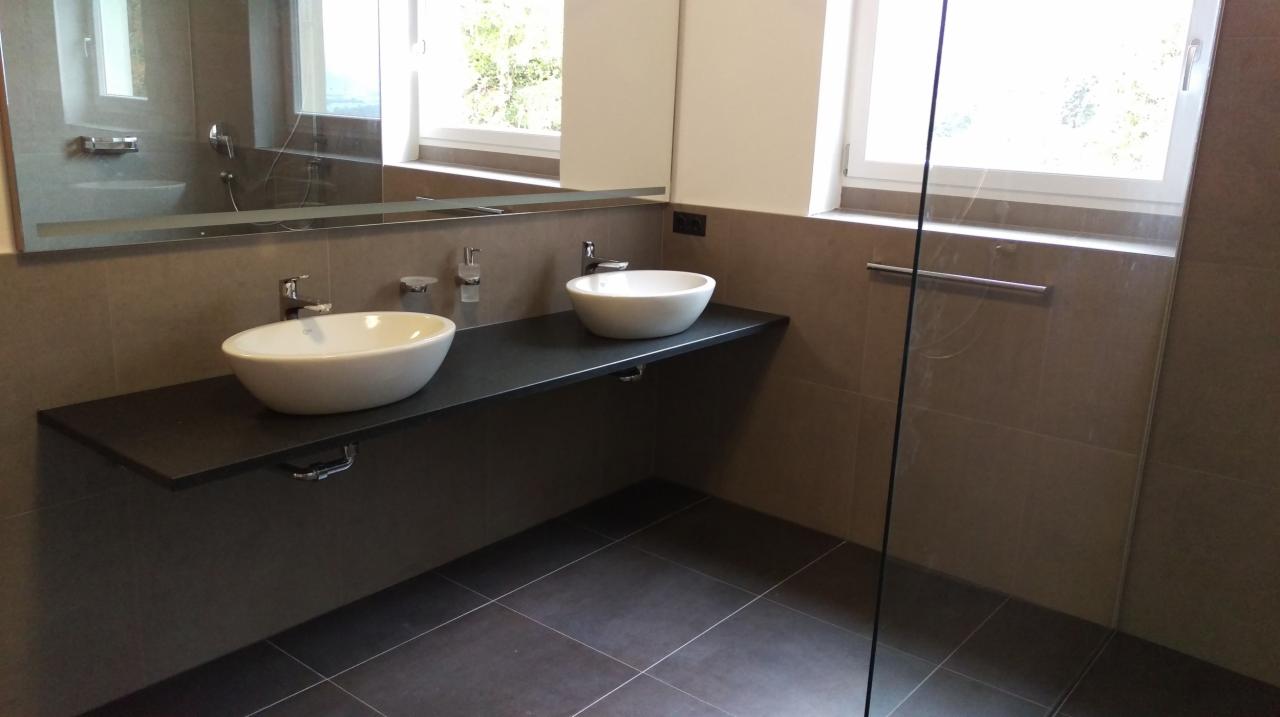 Nowoczesna łazienka z dwoma umywalkami nablatowymi o owalnym kształcie, zamontowanymi na ciemnoszarym blacie, z widocznym lustrem i fragmentem kabiny prysznicowej z przezroczystego szkła.