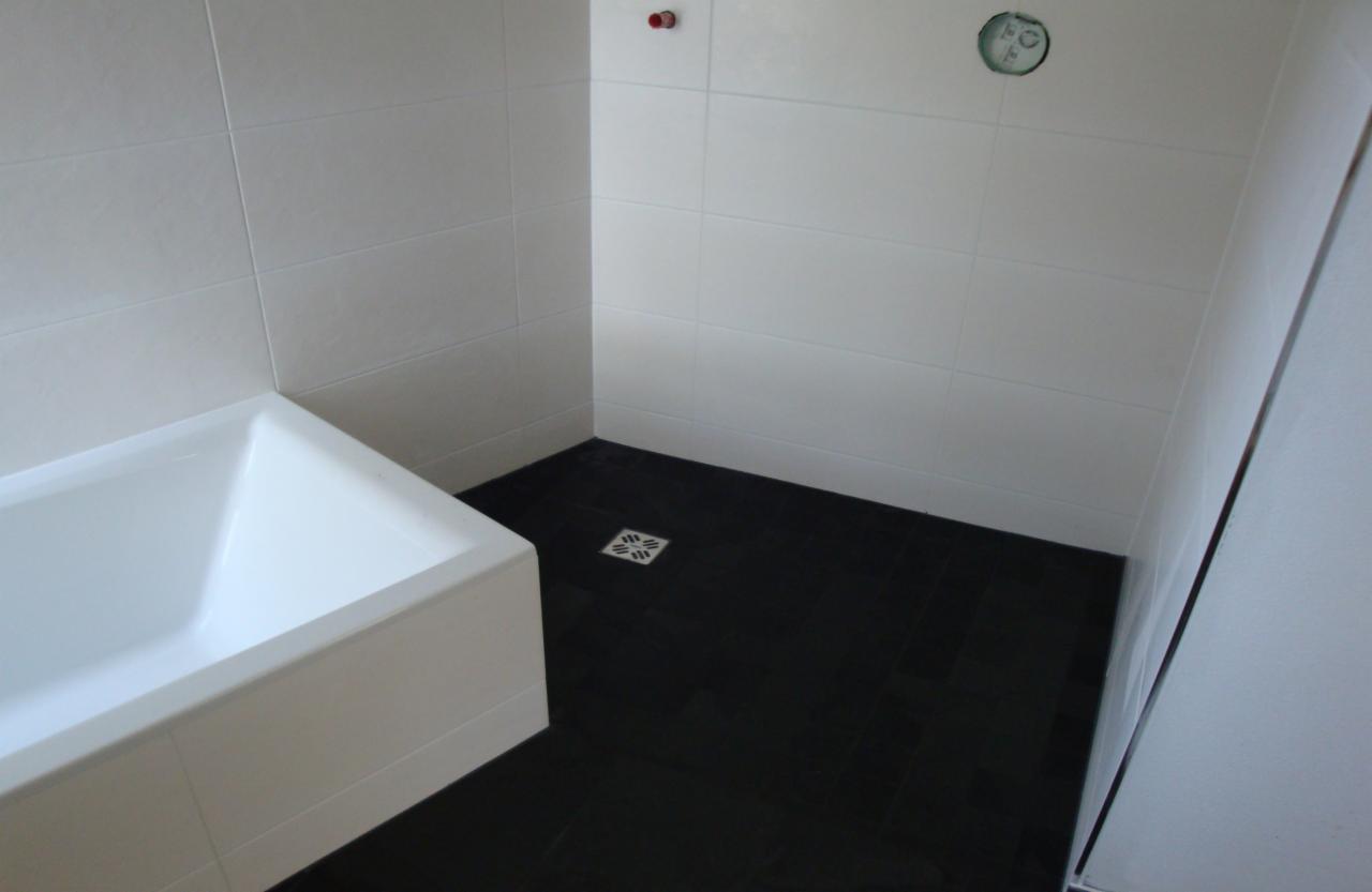 Nowoczesna łazienka z czarną podłogą z płytek i białą wanną, wykończona białymi płytkami ściennymi w połysku, widoczne elementy instalacji.