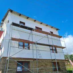 Dom jednorodzinny w trakcie ocieplania, widoczne rusztowanie, przymocowany styropian z różowymi kołkami, dolna część elewacji wykończona kamieniem.