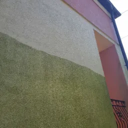 Fragment elewacji budynku z widocznym podziałem na dwie strefy kolorystyczne: dolna część w odcieniach zieleni, górna w beżu, z elementami różu i czerni w tle, sugerujący etap prac związanych...