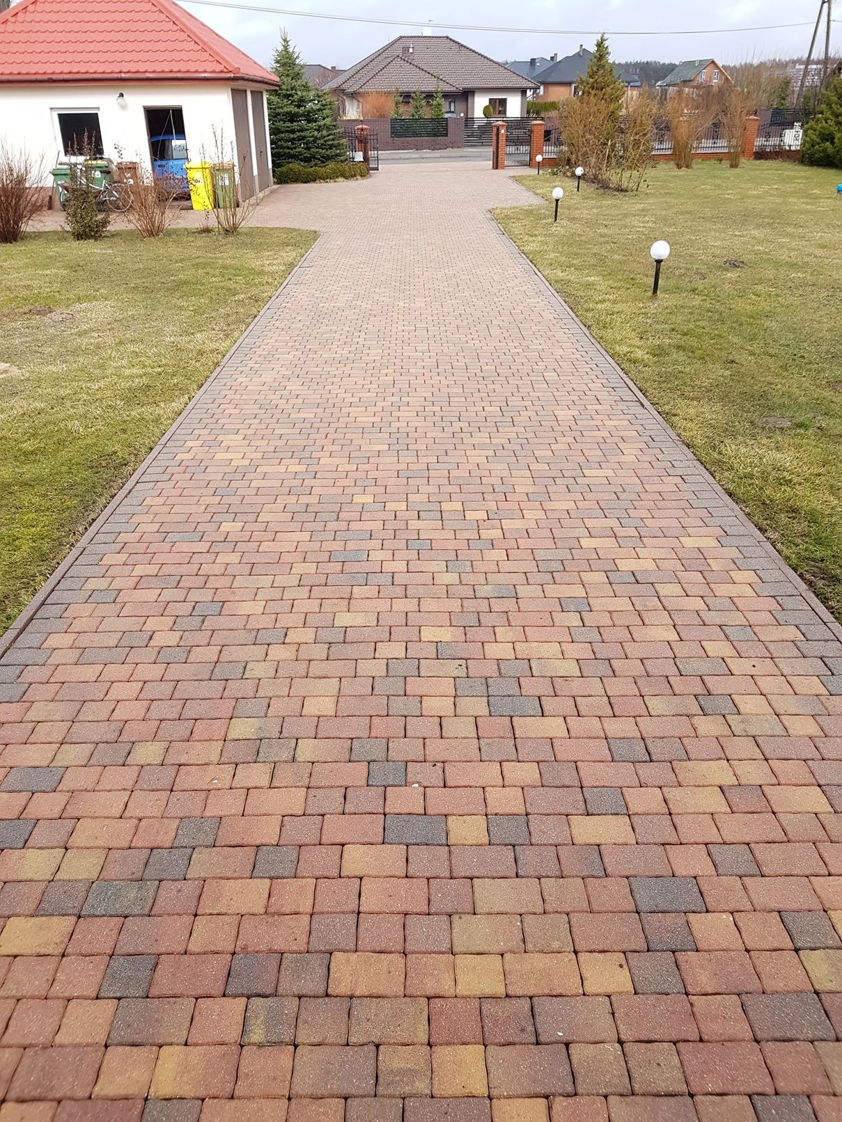 Długa, wyłożona wielobarwną kostką brukową ścieżka prowadząca przez trawnik do domu z czerwonym dachem, oświetlona małymi lampami.