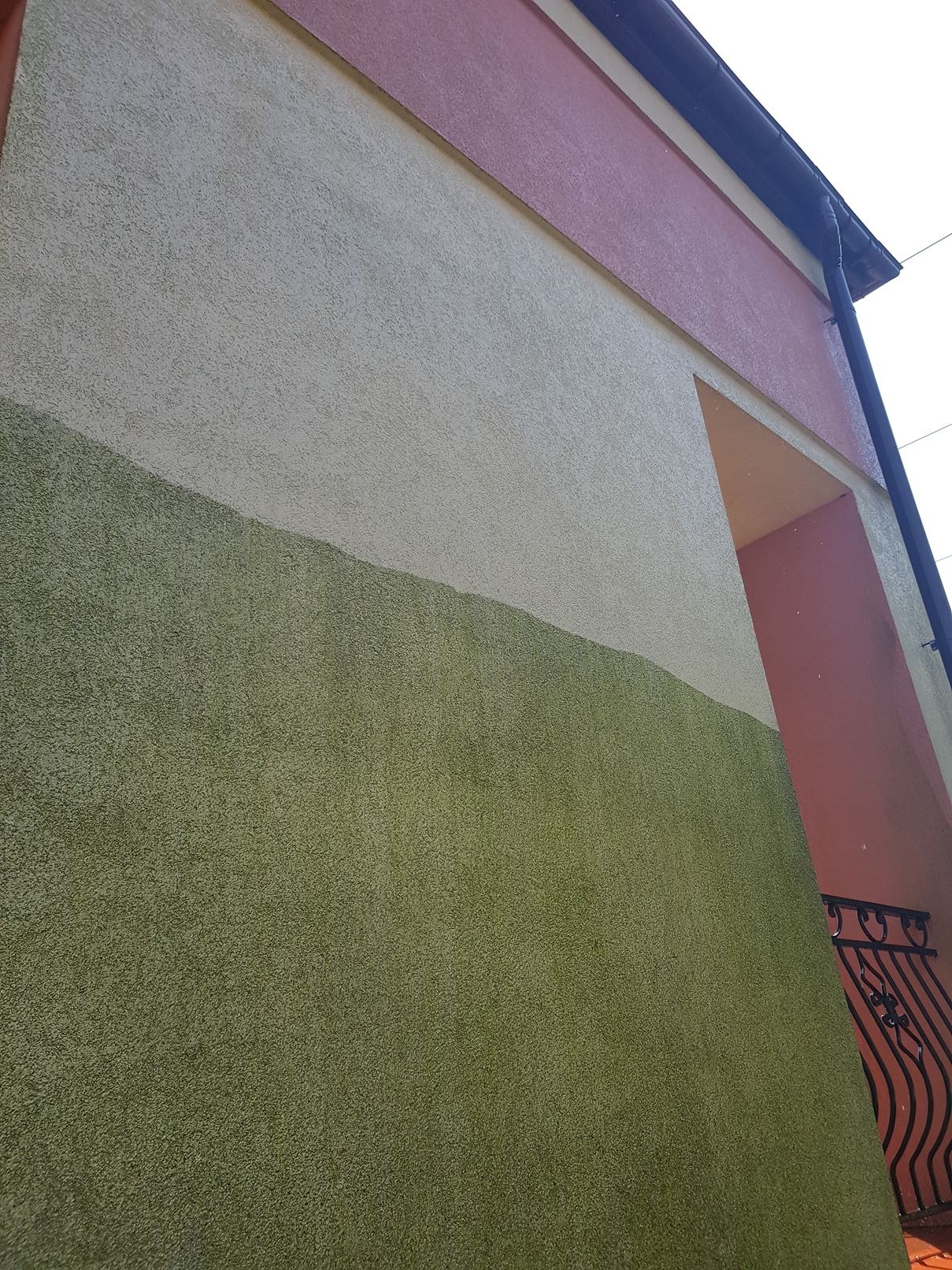 Fragment elewacji budynku z widocznym podziałem na dwie strefy kolorystyczne: dolna część w odcieniach zieleni, górna w beżu, z elementami różu i czerni w tle, sugerujący etap prac związanych...