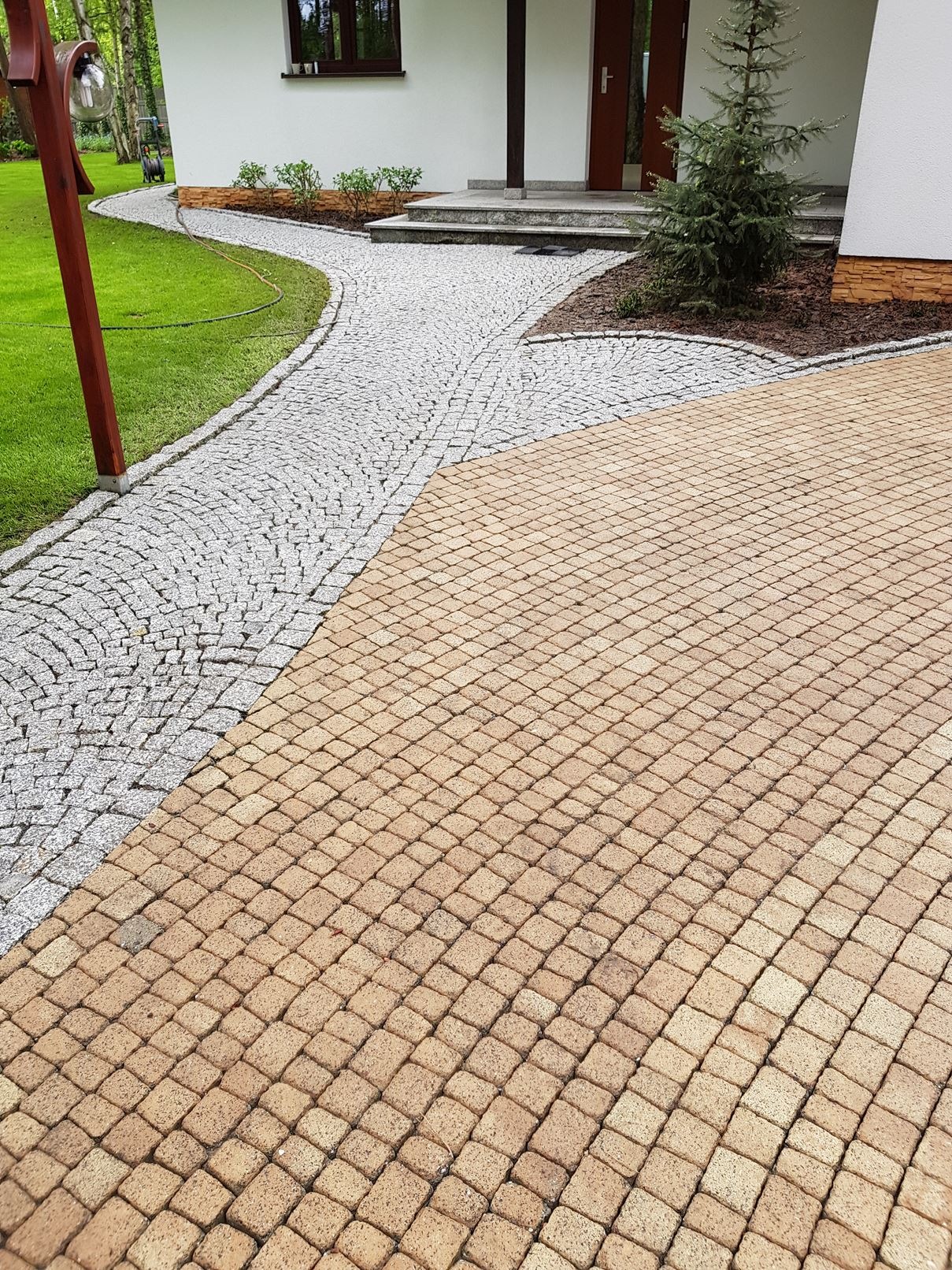 Ułożona z beżowej kostki brukowej i szarego kamienia ścieżka prowadząca do domu z białymi ścianami i małą choinką.