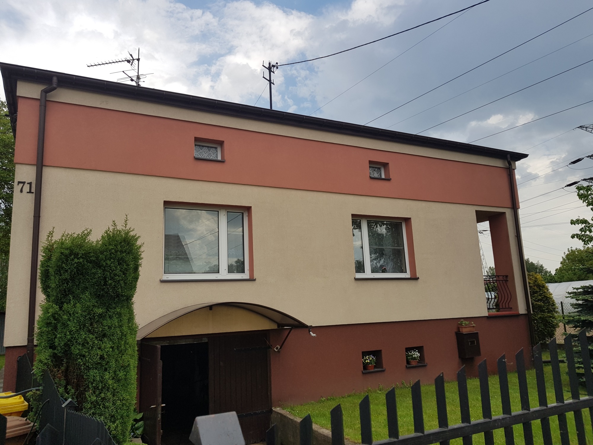 Dwukondygnacyjny dom z beżowo-łososiową elewacją, widoczny zza czarnego metalowego ogrodzenia. Na dachu anteny, a na elewacji małe okienka z firankami. Otwarty garaż w przyziemiu.