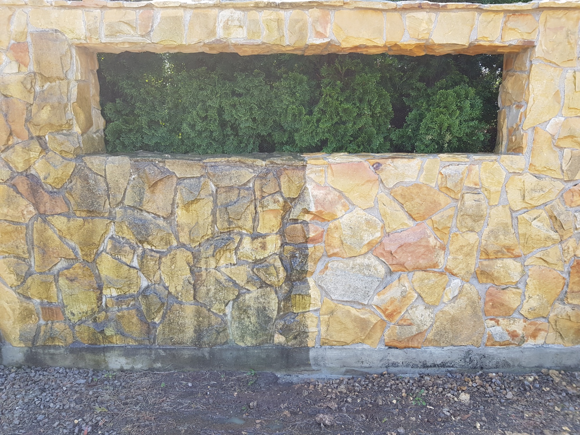 Kamienny mur z oknem, widoczna różnica w czystości połowy muru po zabiegu czyszczenia, zieleń w tle.