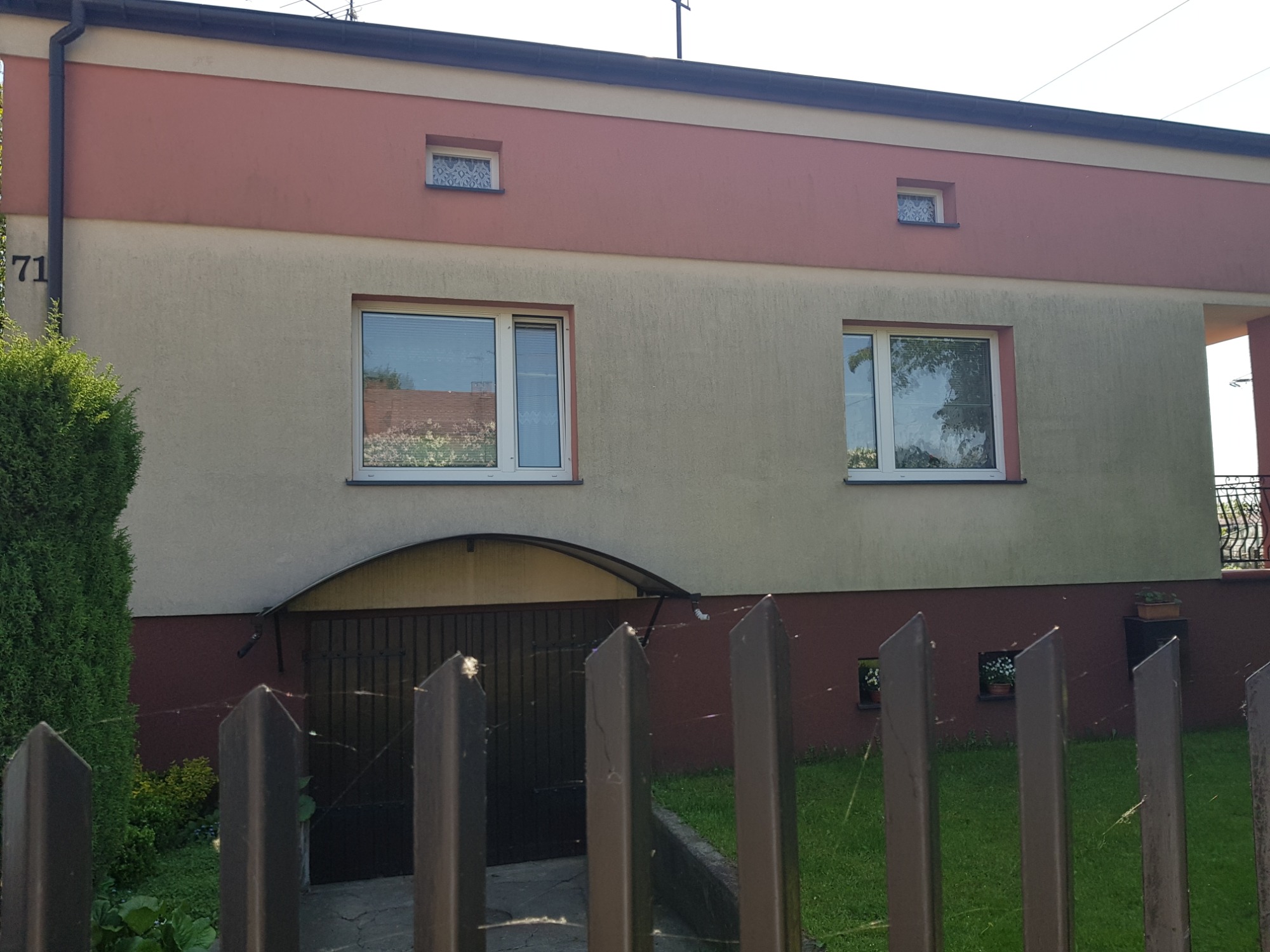 Dwukondygnacyjny dom z elewacją w odcieniach beżu i różu, widziany zza metalowego płotu.