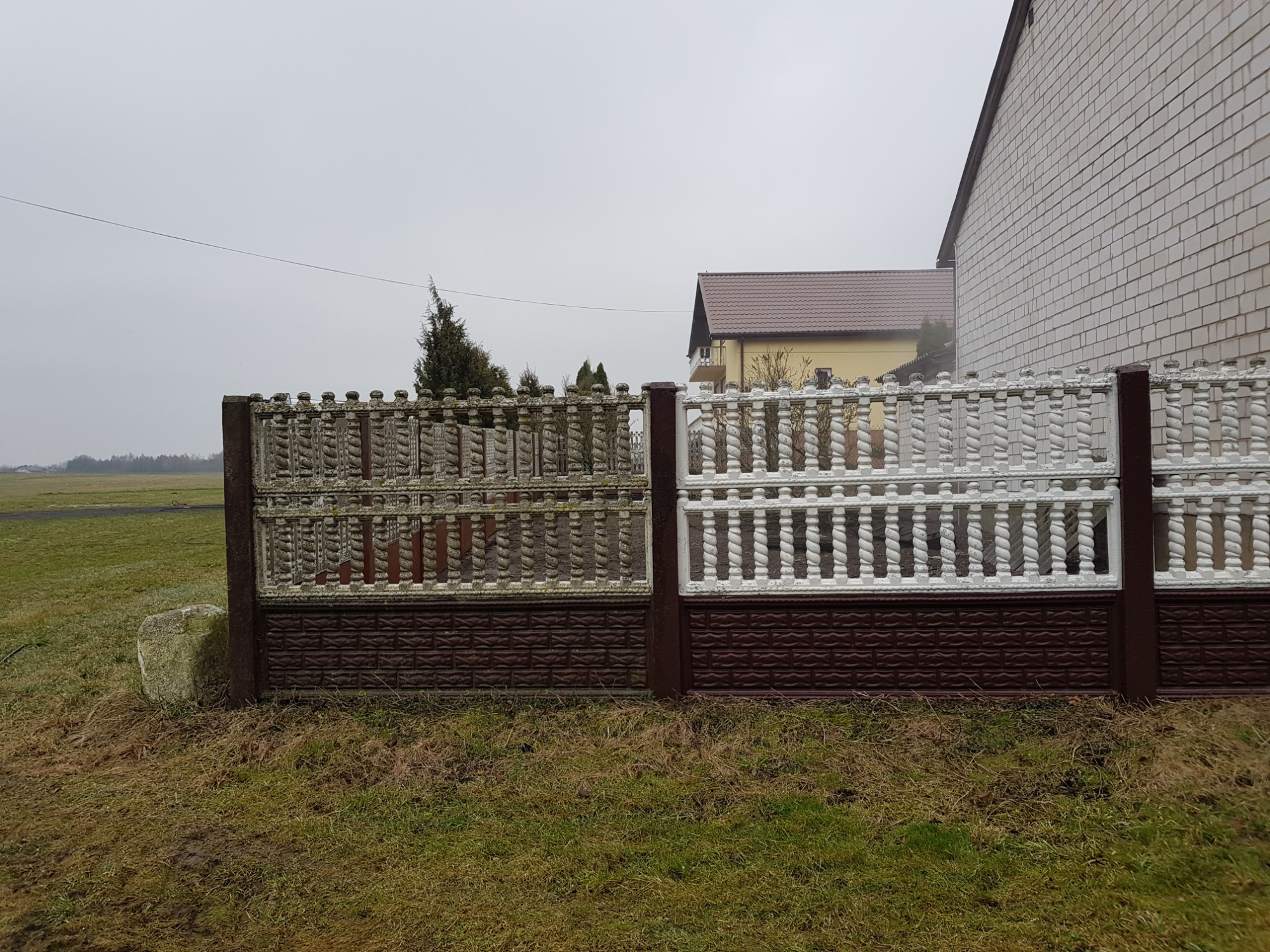 Fragment betonowego ogrodzenia z brązową podstawą, częściowo pomalowany na biało, na tle domu i pochmurnego nieba.