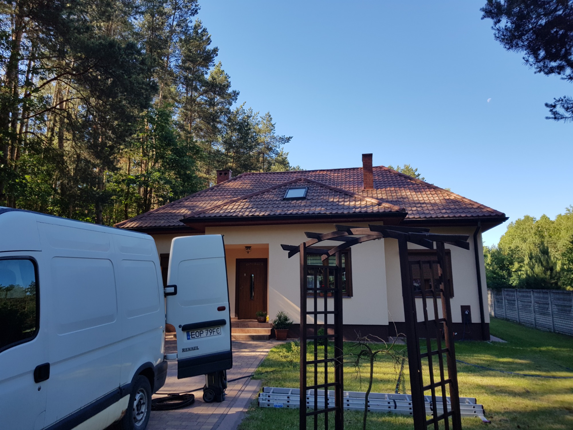 Biały van z otwartymi tylnymi drzwiami stoi przed domem z brązowym dachem i kremową elewacją, widoczna drabina i drewniana pergola.