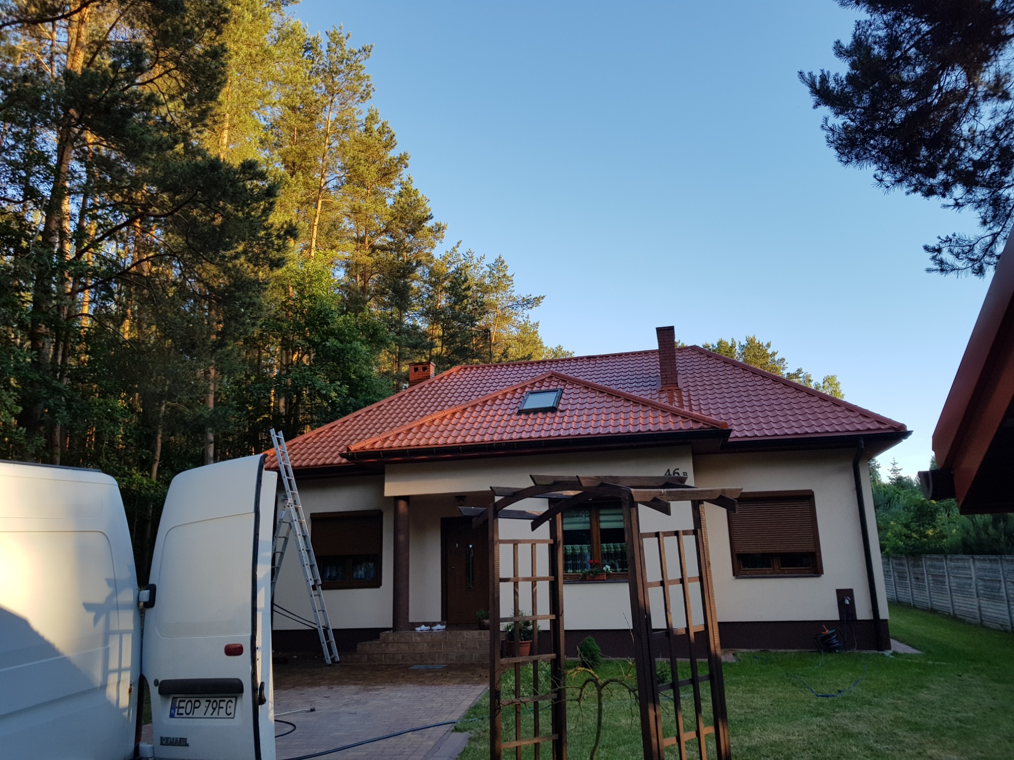 Dom jednorodzinny z czerwoną dachówką, widoczny z boku, z białym vanem i drabiną opartą o rynnę, sugerując prace na wysokości.