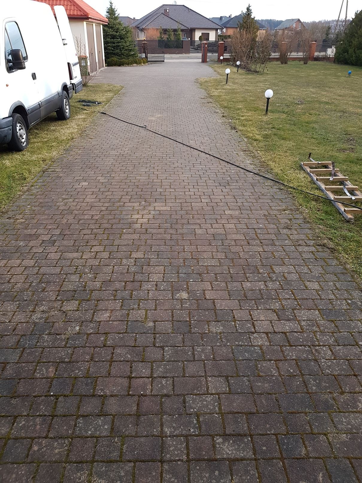 Długa, brudna kostka brukowa prowadząca do domu, z widocznym wężem i drabiną, sugerująca przygotowania do czyszczenia.